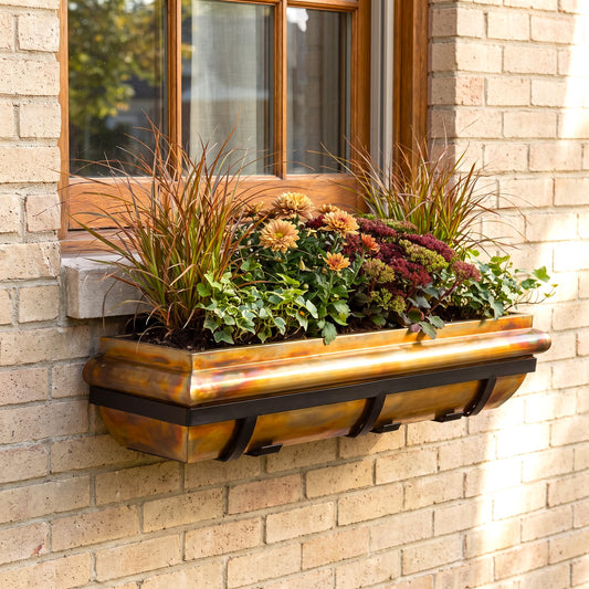 H Potter brand rectangular window box planter mounted on a beige brick wall beneath a wooden window, filled with ornamental grasses, green foliage, and mums in autumn colors.