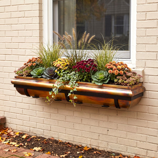H Potter brand rustic copper-toned metal window box planter mounted on a beige brick wall, filled with autumnal flowers and ornamental grasses in front of a white-framed window.