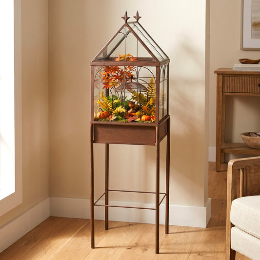 H Potter brand freestanding glass terrarium with a charcoal brown frame and stand, filled with orange and yellow autumn foliage, placed in a real indoor room with wood floors and beige walls.