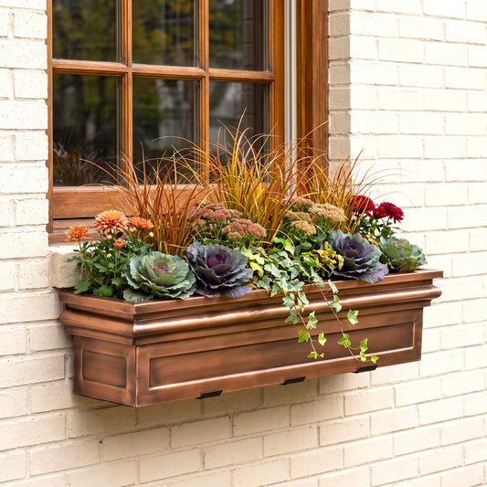A copper-colored metal window box planter mounted on a white brick wall beneath a wooden window, filled with green and purple cabbage, orange flowers, and ornamental grasses.
